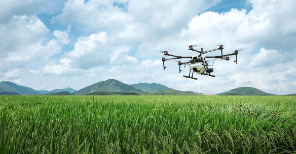 dji agras drone flying over a field of long green grass with mountains in the background and a cloudy blue sky