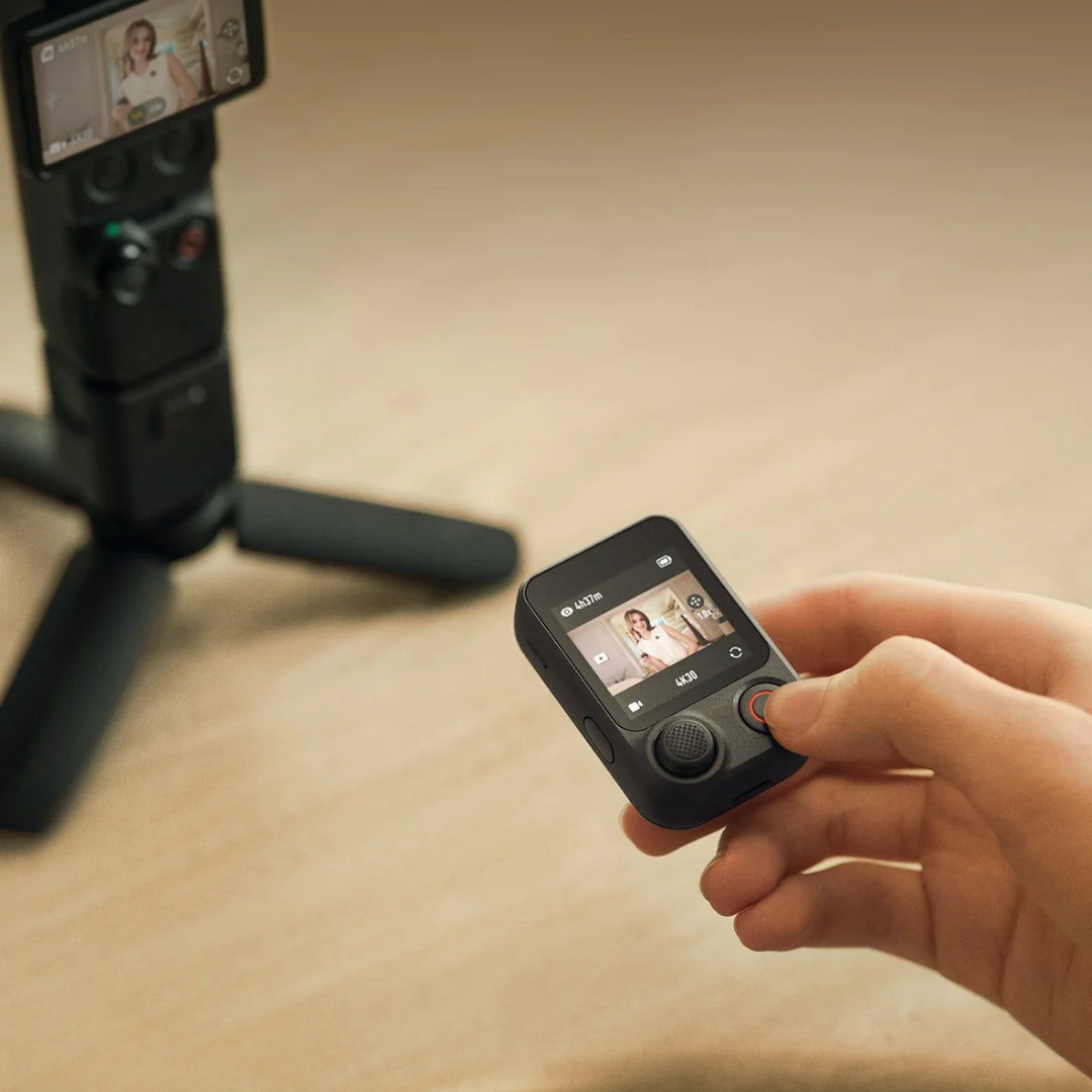 A hand holding a Osmo FrameTap with a screen and thumb wheel, with a DJI Osmo Pocket 4P camera on a tripod blurred in the background on a wooden surface.